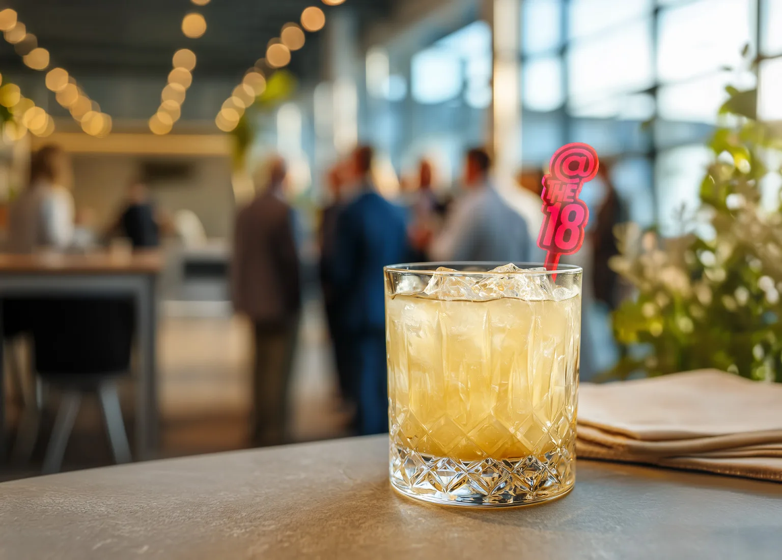 Branded Drink Stirrers for Corporate Events_Experiential Branding Guide. Custom Logo Stirrer in a rocks glass with Corporate Reception in the background.