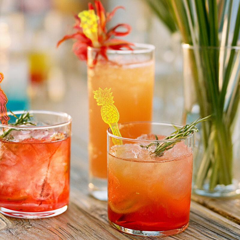 Image of three tropical signature drinks with personalized drink stirrers for a wedding reception on a wood table top with tropical decor. Tiki Stirrer Sets: Hibiscus, Hula Dancer & Pineapple Styles Compared
